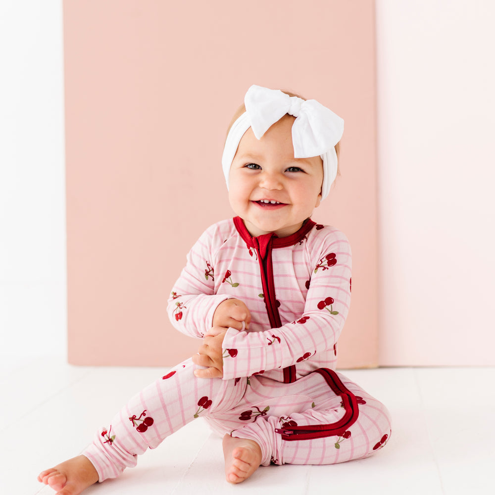 
                      
                        Baby in a pink outfit with red details and a white bow sitting on a white surface.
                      
                    