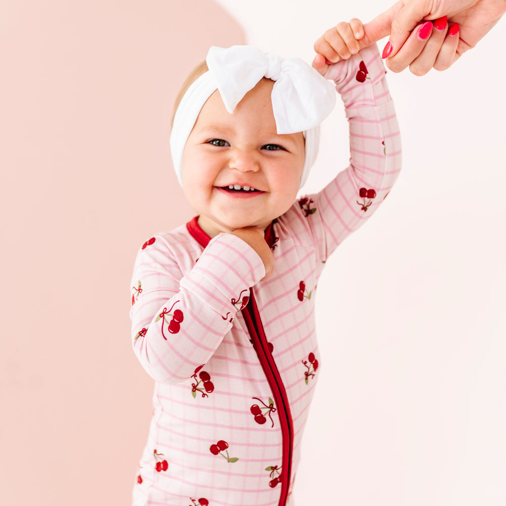 
                      
                        Baby in a pink outfit with red accents and a white headband with a bow, standing against a light background.
                      
                    