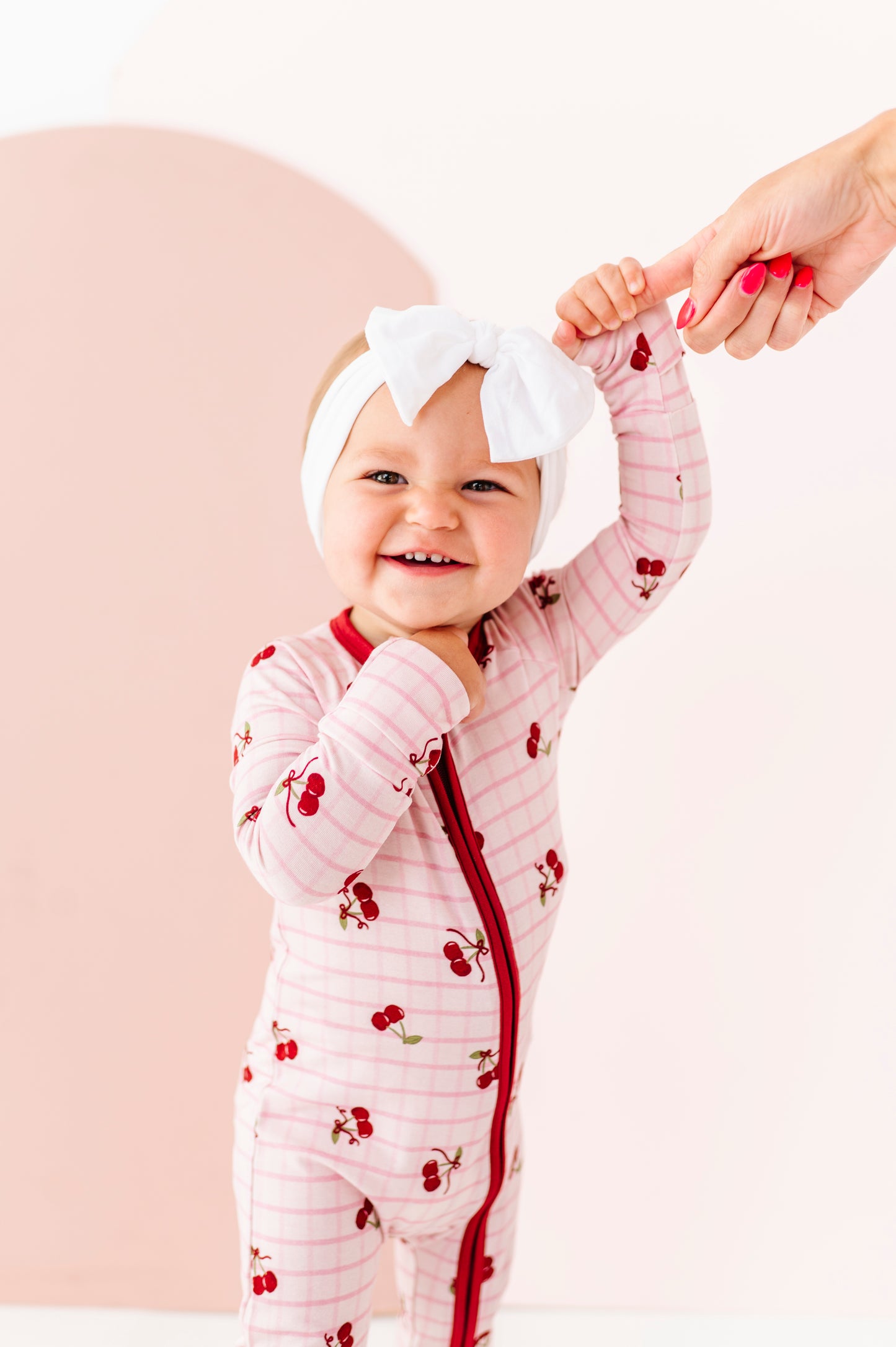 Baby in a pink outfit with red accents and a white headband with a bow, standing against a light background.
