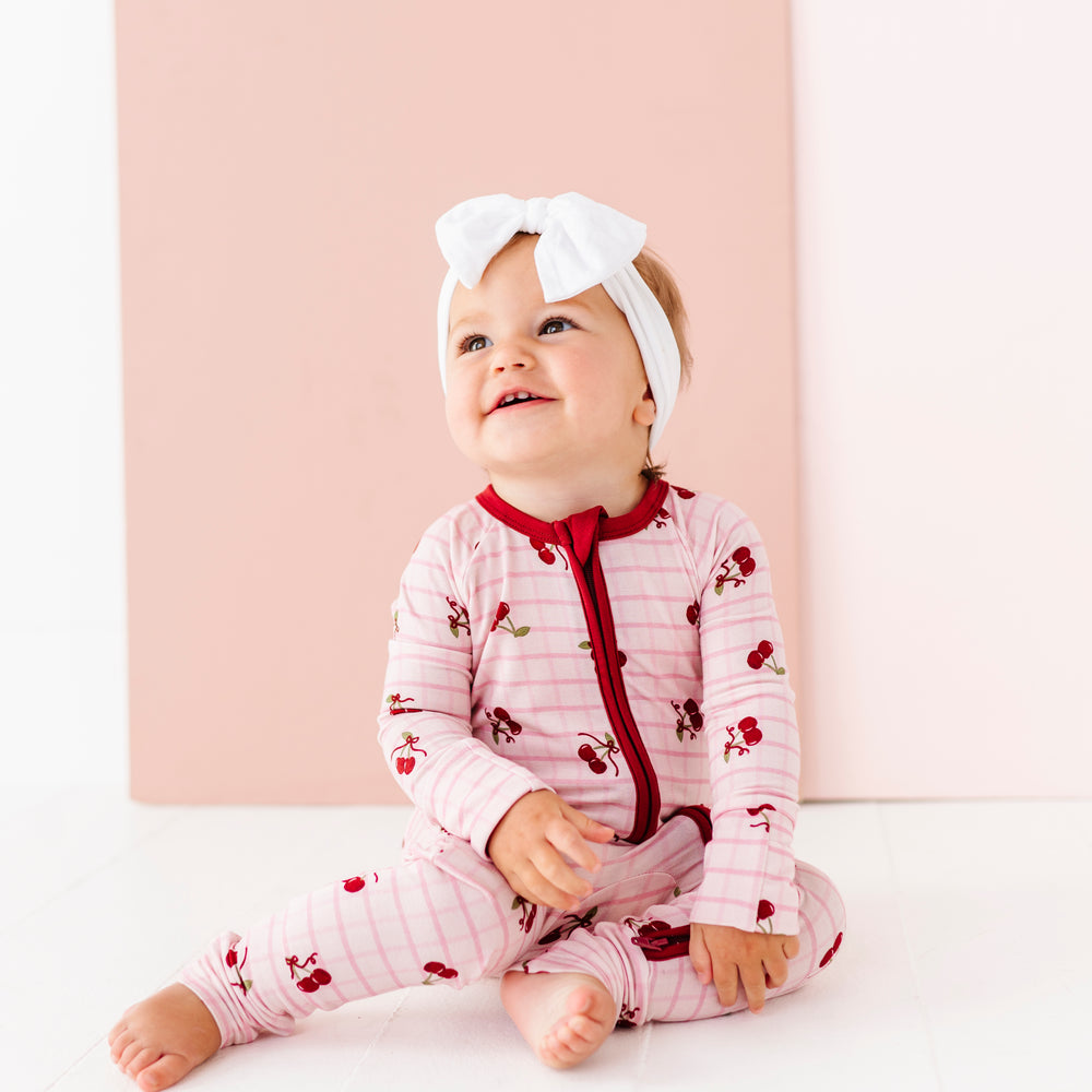 
                      
                        Baby wearing a pink outfit with red accents and a white headband, sitting against a light pink background.
                      
                    