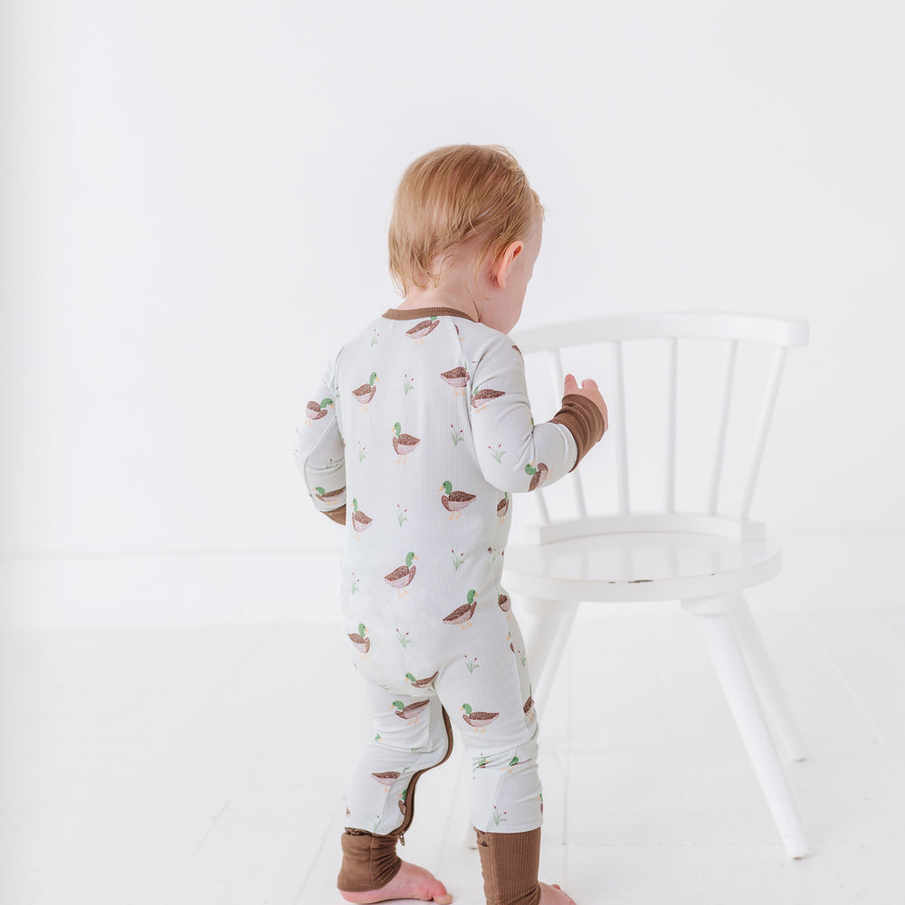 
                      
                        Child wearing a patterned onesie standing next to a white chair on a plain background
                      
                    