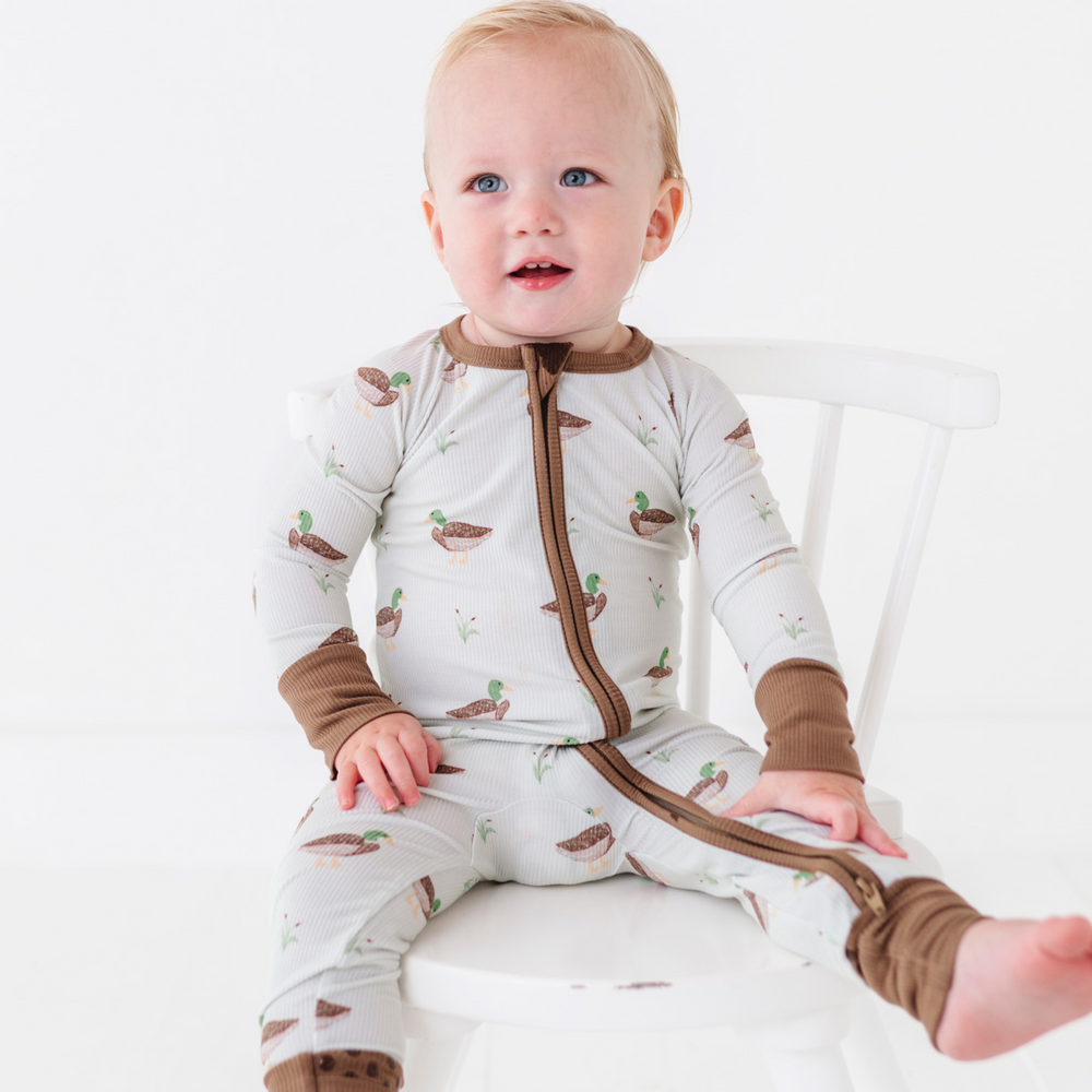 Baby wearing a white onesie with brown accents and bird pattern, sitting on a white chair.
